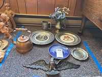 Overview of the lot showing the metal eagle, copper kettle, bicentennial plates, pewter plate, pewter mug, and metal pitcher with faux flowers on carpet next to wooden furniture and wooden dolls.
