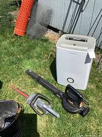 Photo showing Frigidaire humidifier standing on grass outdoors next to a shed wall with two Craftsman electric garden tools lying on the ground