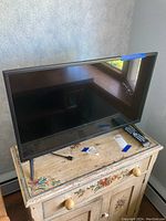 Front view of the 32 inch Insignia TV showing the screen, stand legs, remote, and power cord on a painted wooden cabinet.