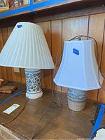 Two ceramic table lamps on a wooden surface, both with attached lamp shades and cords visible, showing overall design and size.