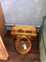 Photo shows a wooden two tier magazine rack with a handle, positioned near a wall and floor. In front of it, there is a handcrafted wooden basket with a woven pattern.