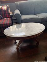 Side view of oval marble top coffee table displaying the wooden curved leg base in a living room setting next to upholstered chairs and sofa.