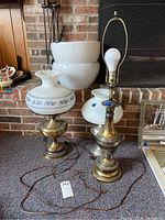 Three metal table lamps with brass finish and different glass shades or no shade shown on carpet in front of brick fireplace.