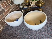 Two matching ceramic planters placed side by side on a carpeted floor against a brick wall, showing one with some dirt residue.