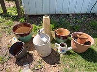 Overview of all planters including glazed ceramic, clay, resin, tall birdbath stand, jug, watering can, and rusty metal pail placed on grass outdoors