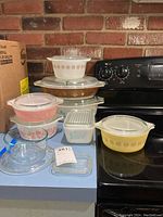 Full assortment of seven Pyrex bowls and dishes with lids displayed on countertop and stove against brick wall.