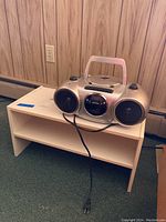 Durabrand silver boombox placed on top of a small storage shelf against a paneled wall; shelf has two open compartments and measures about 24x12x12 inches.