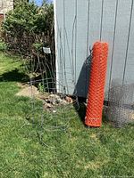 Five metal wire tomato cages standing upright on grass near shed, with visible rust and wear. Next to cages are rolls of orange plastic mesh fencing and black wire mesh fencing leaning against shed wall.