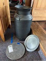 Vintage metal milk can standing upright next to a pewter plate and embossed round trivet on a carpeted floor