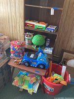 Wide view showing assorted toys placed on and near a table, including a blue Fisher Price ride-on car, a green tree toy, vintage board games, and a bucket of plastic pieces.