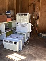 Four window unit air conditioners stacked against wooden wall and shelving, cables visible, various control panel designs.