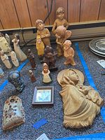 Wide view of the lot showing multiple wooden figurines of children and animals, resin and stone figurines, pottery wall pocket, and framed quote all positioned on a carpeted surface lined with blue tape.