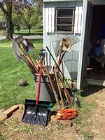 Assorted yard tools including snow shovel, spade shovels, rakes, axes, sledge hammer, broom, heavy-duty extension cord, and a large garbage can placed outdoors next to a shed.