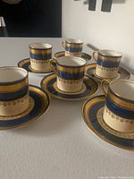 Photo showing 6 Aynsley demi tasse coffee cups arranged with their matching saucers, showing the cream ground with wide blue and gold detailing.