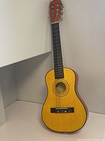 Front view of small-sized children's acoustic guitar standing against a wall showing the full instrument and its natural wood finish.