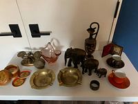 Overall view of antique decorative objects including painted coasters, bronze scale, pink glass vase, carved elephants, porcelain cups, and assorted metal pieces on white surface.