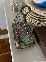 Full item shot showing the silver plated toast holder with a handle and a beaded edge base sitting on a surface near stacked plates.