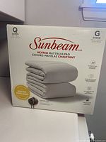 Front of box showing brand Sunbeam, product type heated mattress pad in queen size, image of folded quilted pad.