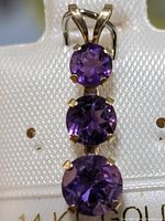 Close-up image of a 14K gold pendant featuring three round amethyst gemstones in prong settings arranged vertically.