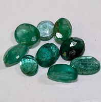 Close-up of ten loose genuine emerald gemstones varying in shape and color, laid on a flat white surface.