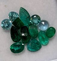 Photo showing a cluster of 13 variously shaped and sized emerald gemstones on a light background.