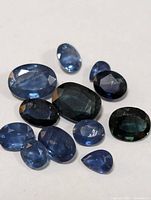 Photo showing 11 loose sapphire gemstones with various blue and teal colors, faceted cuts, oval and pear shapes, and visible inclusions.
