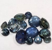 Close-up photo showing a collection of twelve loose faceted sapphires in various shades of blue and cuts arranged on a white background.
