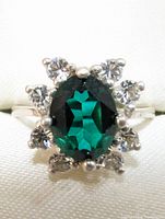 Close-up of silver ring with green oval central gemstone and surrounding clear stones on silver band.