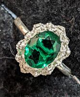 Close-up of silver ring with large faceted oval green gemstone and textured silver halo on black background.