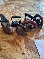 Photo showing 6 antique small irons arranged on wood surface, diverse handle styles and heavy rust.