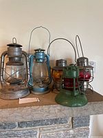 Photo showing five antique lanterns placed on a mantel with varying sizes, metal frames and glass globes in silver, blue, amber, red, and green colors.