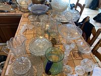 Photo showing a variety of clear and some tinted cut glass items including large pitchers, covered dishes, and bowls arranged on a tiled table surface.