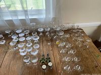 Wide view showing approximately 60 assorted clear glass stemmed glasses arranged on wooden table along with a glass floral base holder containing salt and pepper shakers.