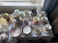 Wide view of assorted teacups, saucers, sugar pots and creamers on a table near window, showing different styles and colors.