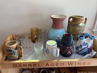 Photo of assorted glasses, mugs, and ceramic vases on wooden tray labelled 'Barrel Aged Wines'. Includes clear glass tumblers, a brown and blue geometric vase, a tan and turquoise small vase, two small mugs, and an amber glass.