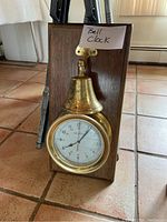Clock mounted on a wood plaque with a brass round clock face and brass bell above it, showing front view with the clock closed.