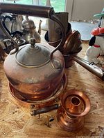 Photo showing a copper teapot with a wooden handle, a small copper pot, and a copper candlestick placed on a wooden table.