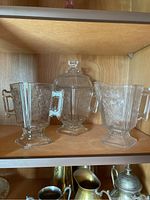 Three clear glassware pieces on a wooden shelf, featuring cut flower designs and handled pitchers alongside a lidded container.