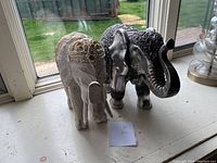 Set of two decorative elephant figurines shown from a side and rear angle by a window, highlighting the silver and black elephant in the background and the white and gold elephant in the foreground.
