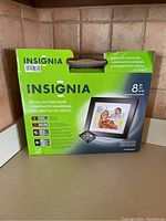 Photo of the boxed Insignia 8-inch digital picture frame on a kitchen counter, showing front and side of box with branding and product information.