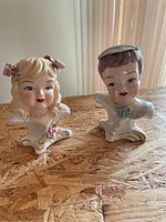 Two porcelain figurines shaped as a girl's and boy's head placed on a wooden surface with a curtain background
