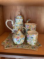 Photo shows a 5-piece porcelain tea set with floral pattern and gold accents on a wooden shelf.