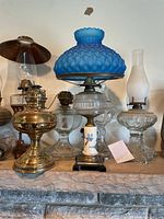 Photo of six different oil lamps made of brass, glass, porcelain with variations in chimney and shade designs.