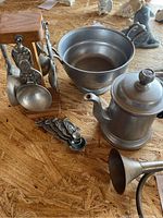 Photo displaying pewter pieces including a small bowl, typical measuring spoons on a wooden stand, and a teapot with lid