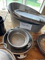 Photo showing several metal roasting pans stacked, including a large oval roasting pan with lid and multiple round pans with handles.