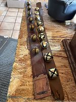 Two brown leather strips with attached brass sleigh bells placed side by side on a wooden surface.