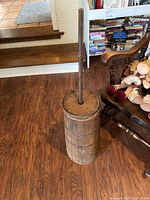 Full view of the vintage wooden butter churn placed on a wooden floor next to a chair and bookshelf.