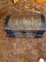 Closed view of a vintage dome-top wooden chest with black and bronze decorative metal bands on a brown carpet.