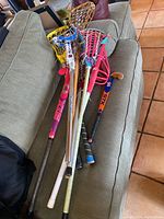 Photo showing seven youth lacrosse and field hockey sticks laid on couch, including plastic head lacrosse sticks in yellow, red, brown and wooden traditional lacrosse sticks, plus field hockey sticks in black and orange colors.