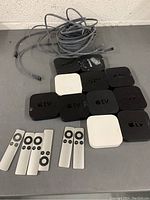 Photo of 9 Apple TV units stacked on floor with 7 Apple TV remotes and assorted cables coiled